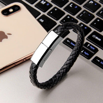 Wearable smartphone charging cable bracelet with a polished silver rectangular magnetic clasp, displayed diagonally on a light gray surface next to a smartphone and a laptop keyboard.