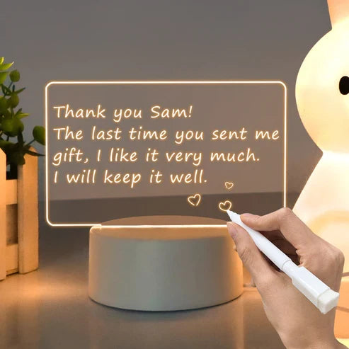 LED acrylic message board with light-up writing with a message being written on it by a hand, with a plant and lamp in the background.