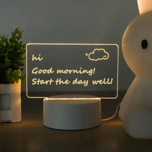 LED acrylic message board with light-up writing  on a round base, displaying 'hi Good morning! Start the day well!' with a small cloud drawing. The lamp sits on a dark tabletop next to a potted plant and a white bunny-shaped lamp.