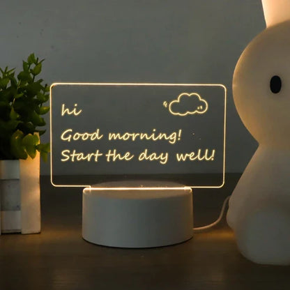 LED acrylic message board with light-up writing  on a round base, displaying 'hi Good morning! Start the day well!' with a small cloud drawing. The lamp sits on a dark tabletop next to a potted plant and a white bunny-shaped lamp.