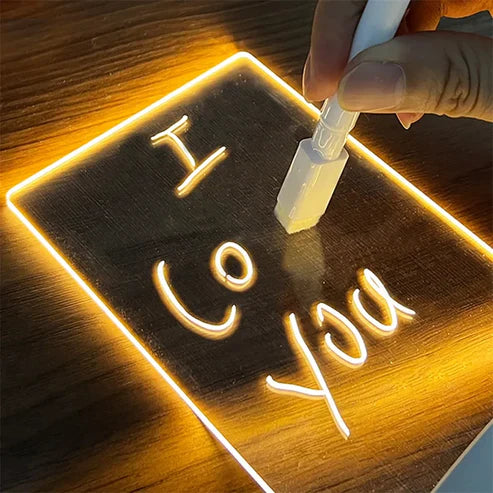 A hand uses a white marker to write 'I Co you' on a LED acrylic message board with light-up writing that illuminates the wood surface with a warm yellow light.